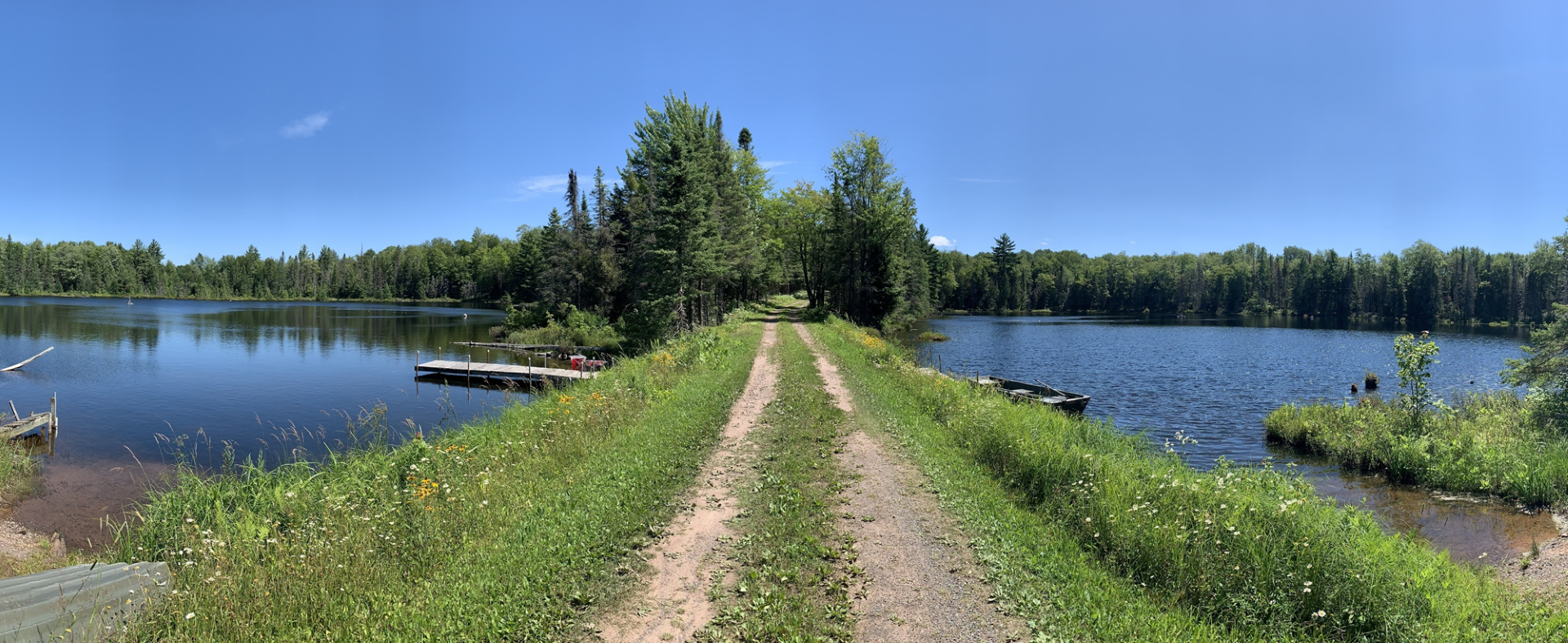 There are two lakes on either side of a one car path between the two, with grass growing on the dam; it's a sunny, blue sky day with tree line in the background.