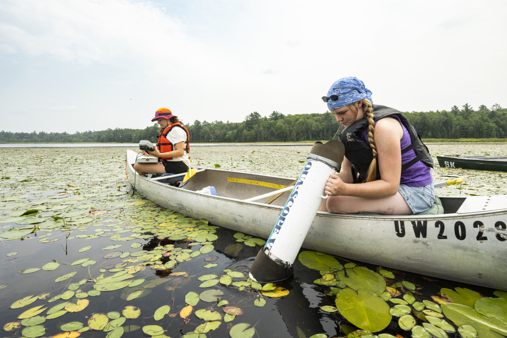 Two students sitting on opposite ends of canoe in lake surrounded by trees shoreline. One is taking notes and the other is looking through large scope into the water.