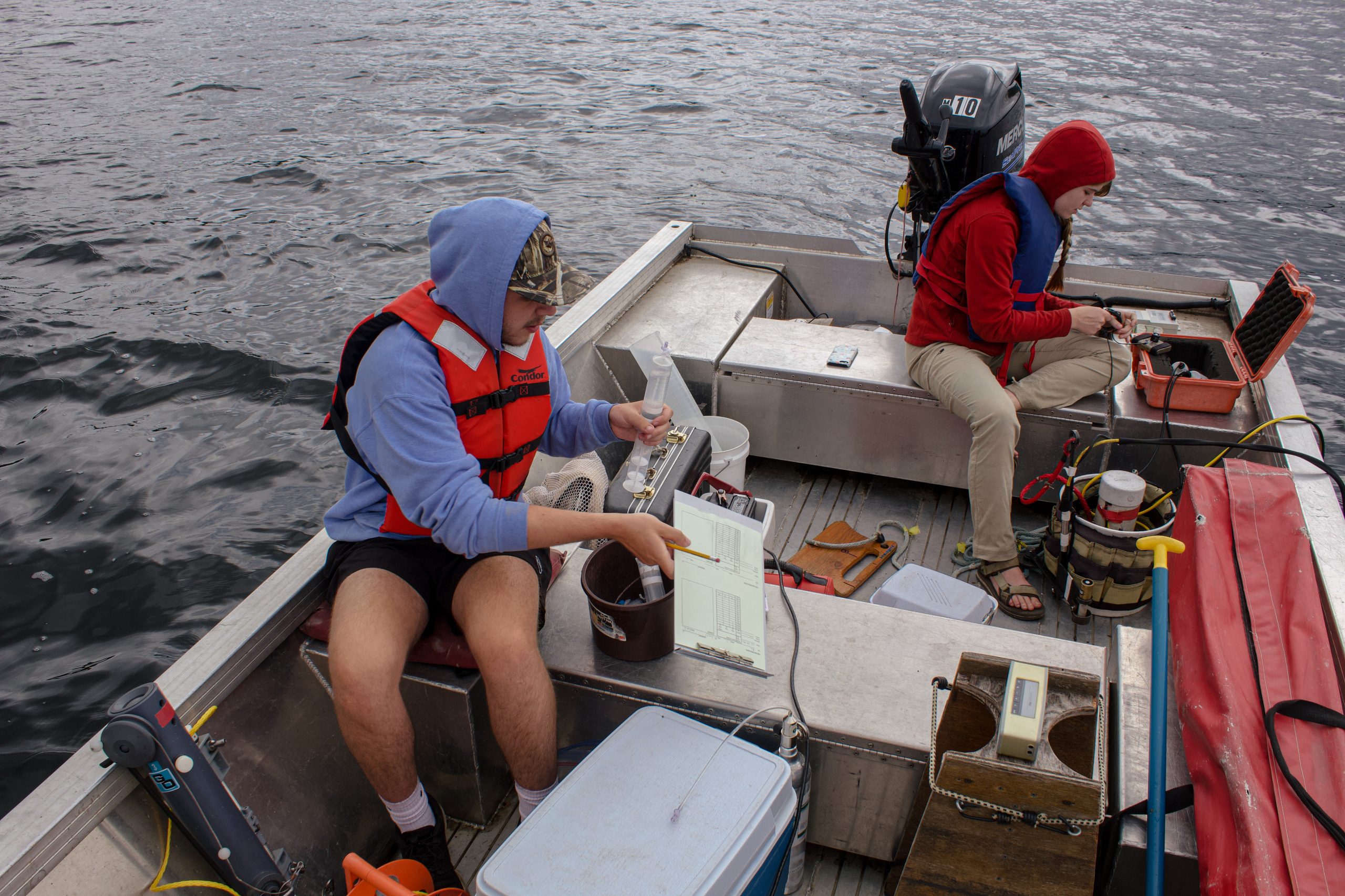 There are two people wearing hoodies, sitting in john boat recording data collected from lake.