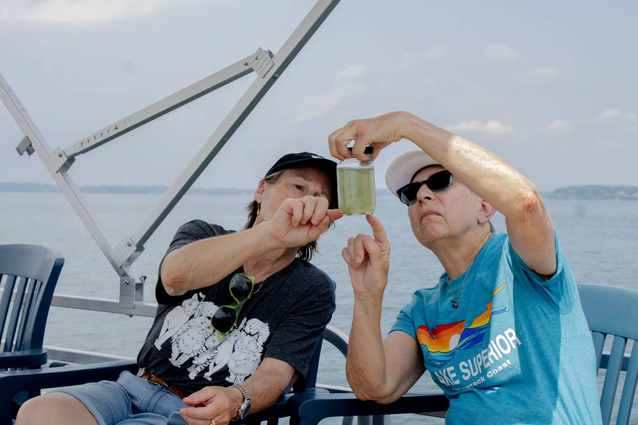 Two people are sitting on a pontoon boat during Open House at Hasler Lab. They are wearing tshirts and shorts and peering at and pointing to zooplankton in bottle of water from Lake Mendota.