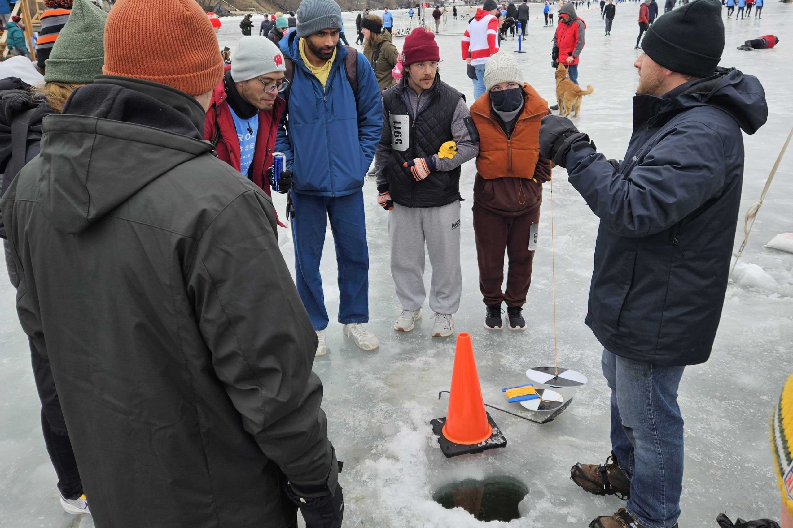 Group of people are standing on frozen Lake Mendota around a hole in the ice as CFL grad student explains winter limnology