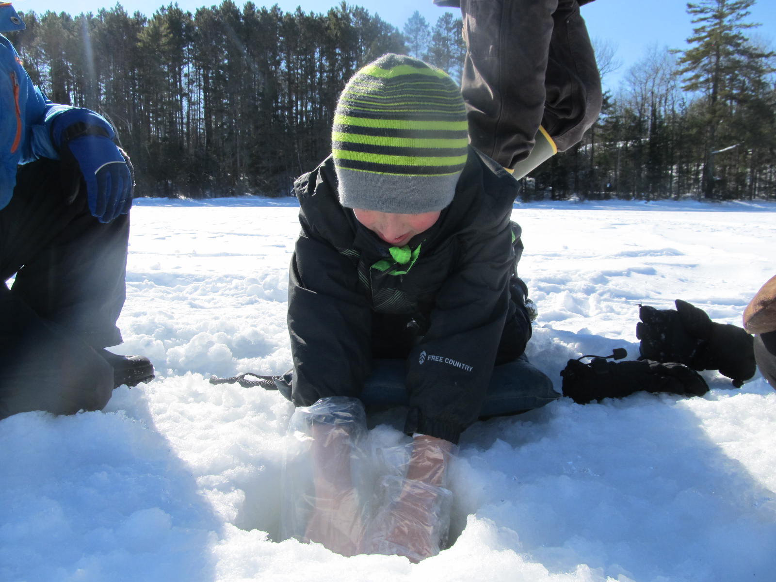 A child is dressed in winter clothing and is peering into a hole in the snow covered lake ice.
