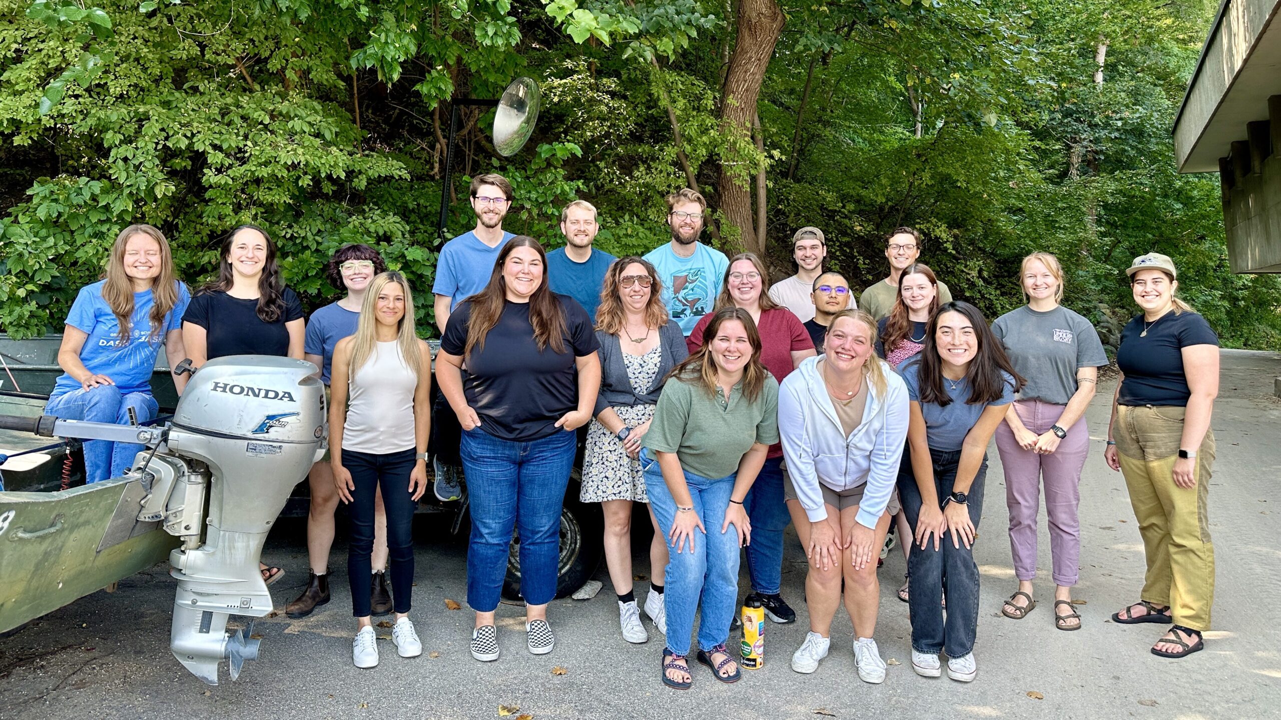 Photo taken in 2024 of group of graduate students standing at side of Hasler Lab with trees in background 